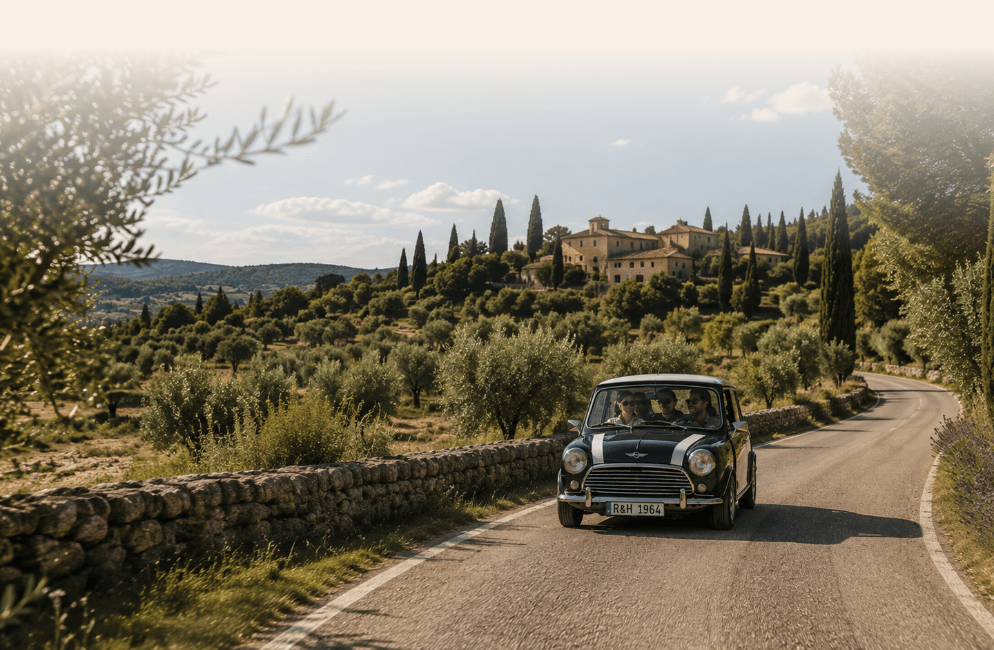 Une Austin Mini vert anglais roulant sur une route de campagne bordée d’oliviers et de murets de pierres sèches, avec un hameau perché en arrière-plan, dans la lumière du jour.