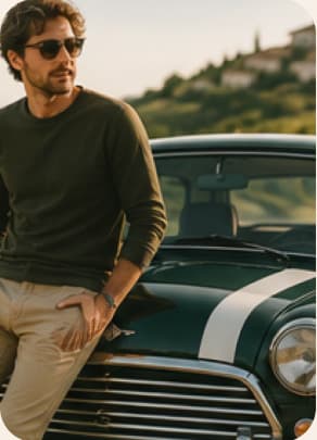 Un homme en pull sombre et lunettes de soleil, adossé au capot d’une Mini, face aux collines.